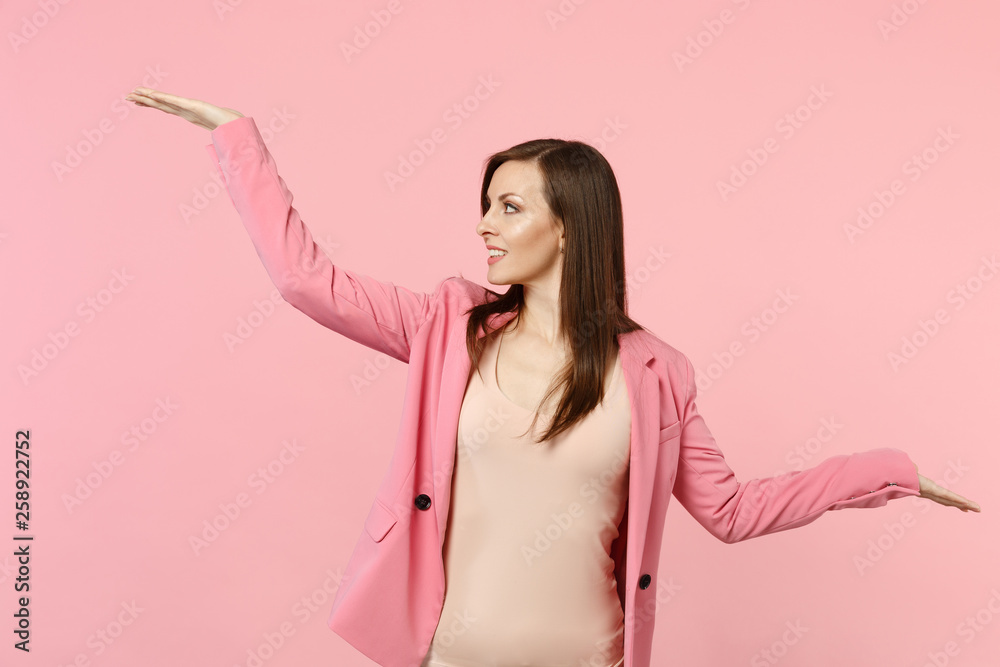 Portrait of smiling woman wearing jacket looking, spreading and pointing hands aside isolated on pastel pink wall background in studio. People sincere emotions, lifestyle concept. Mock up copy space. Portrait of smiling woman wearing jacket looking, spreading and pointing hands aside isolated on pastel pink wall background in studio. People sincere emotions, lifestyle concept. Mock up copy space.