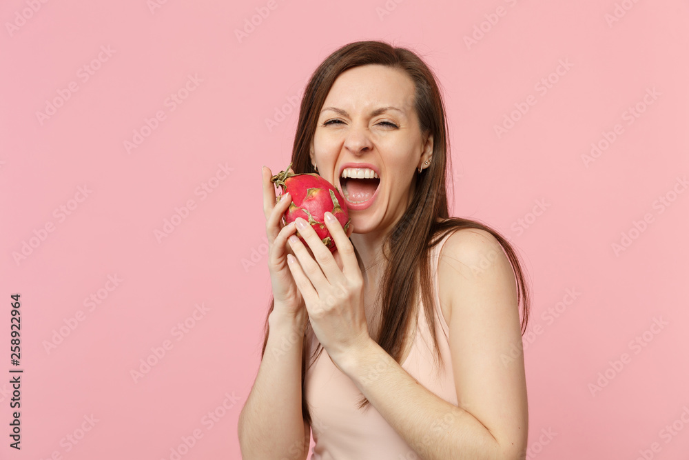 Crazy young woman in light clothes hold, biting fresh ripe pitahaya, dragon fruit isolated on pink pastel wall background in studio. People vivid lifestyle, relax vacation concept. Mock up copy space. Crazy young woman in light clothes hold, biting fresh ripe pitahaya, dragon fruit isolated on pink pastel wall background in studio. People vivid lifestyle, relax vacation concept. Mock up copy space.