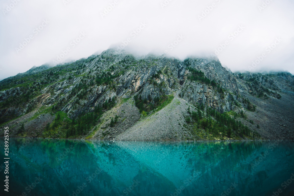 Giant cloud above rocky mountainside with trees in fog. Amazing mountain lake. Mountain range under cloudy sky. Wonderful rocks in mist. Morning landscape of highland nature. Low clouds in mountains.