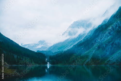 Ghostly forest near mountain lake in early morning. Mountain creek flows into lake. Ripple on smooth water surface. Low clouds. Dark calm atmospheric misty woodland landscape. Tranquil atmosphere. © Daniil