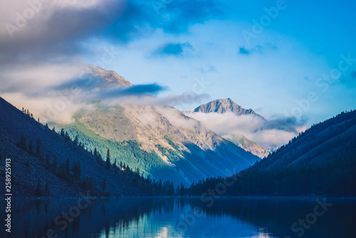 Fototapeta Naklejka Na Ścianę i Meble -  Amazing blue silhouettes of mountains under blue cloudy sky. Beautiful ripples on water of mountain lake. Low clouds before mountain ridge. Wonderful highland landscape. Picturesque mountainscape.