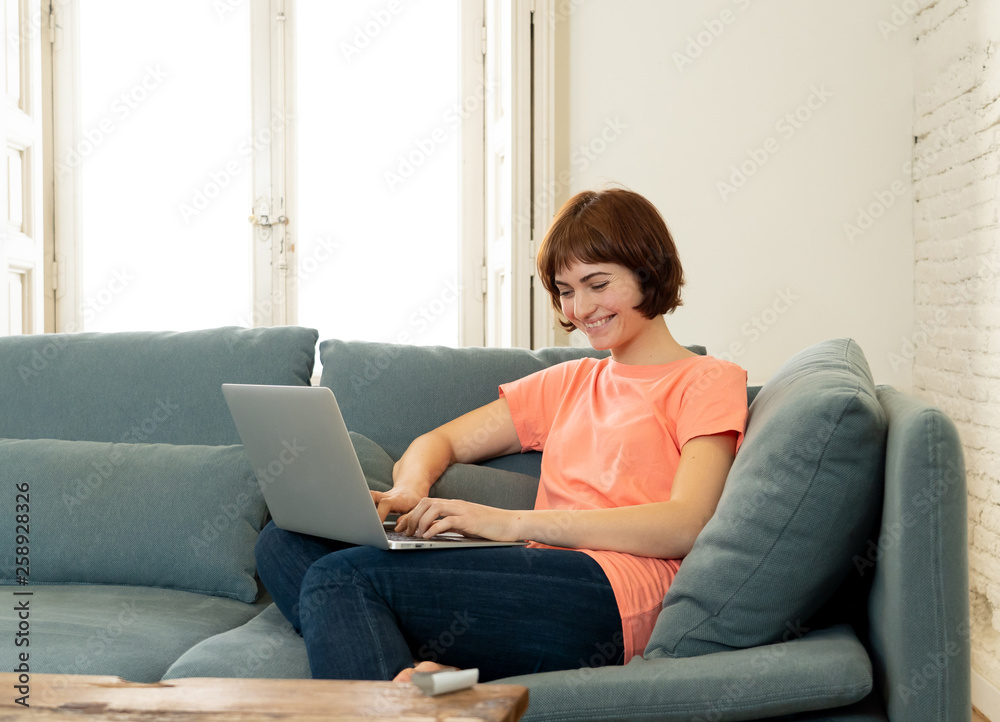 Happy young woman working on her laptop sitting on the sofa. In education and working concept