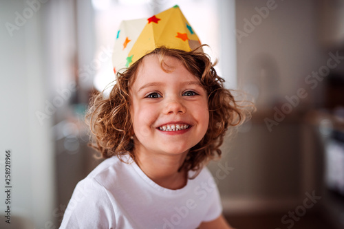 A small girl with a paper crown at home, looking at camera.