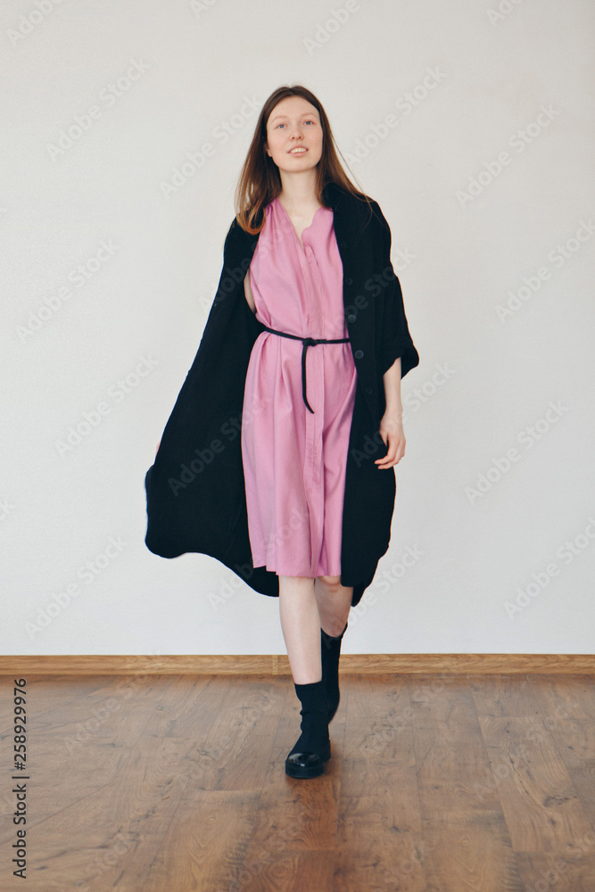 Beautiful girl without makeup in pink dress with black belt under black linen coat in black shoes on home interior background Beautiful girl without makeup in pink dress with black belt under black linen coat in black shoes on home interior background