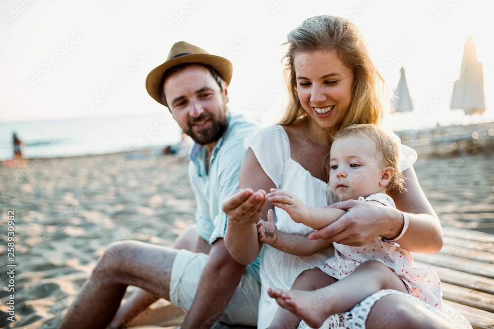 A family with a toddler girl sitting on sand beach on summer holiday. - obrazy, fototapety, plakaty 