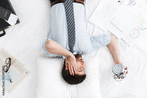 Shocked handsome young businessman in the morning in bed lies sleeping holding alarm clock.
