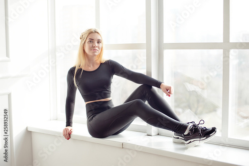 Young slim woman with an athletic body long blonde hair wearing in black sports sportswear top and leggings standing in bright yoga room with big panoramic window preparing before training health life