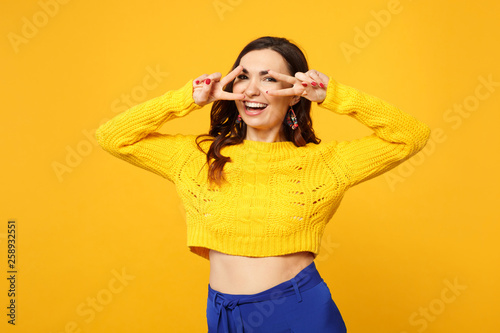 Portrait of laughing young woman in sweater, blue trousers looking camera showing victory sign isolated on yellow orange wall background. People sincere emotions lifestyle concept. Mock up copy space.