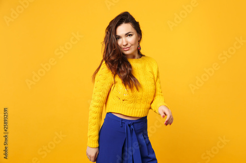 Portrait of smiling young woman in sweater, blue trousers standing, looking camera isolated on yellow orange wall background in studio. People sincere emotions, lifestyle concept. Mock up copy space.