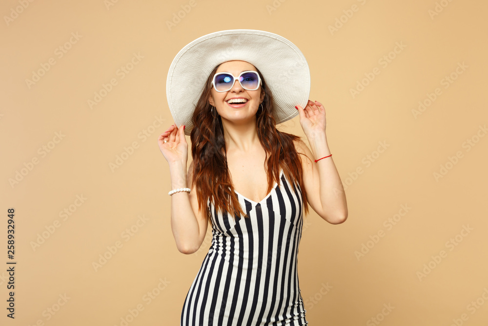 Portrait of smiling young woman in black and white striped dress, hat and sunglasses looking camera isolated on pastel beige background. People sincere emotions, lifestyle concept. Mock up copy space. Portrait of smiling young woman in black and white striped dress, hat and sunglasses looking camera isolated on pastel beige background. People sincere emotions, lifestyle concept. Mock up copy space.