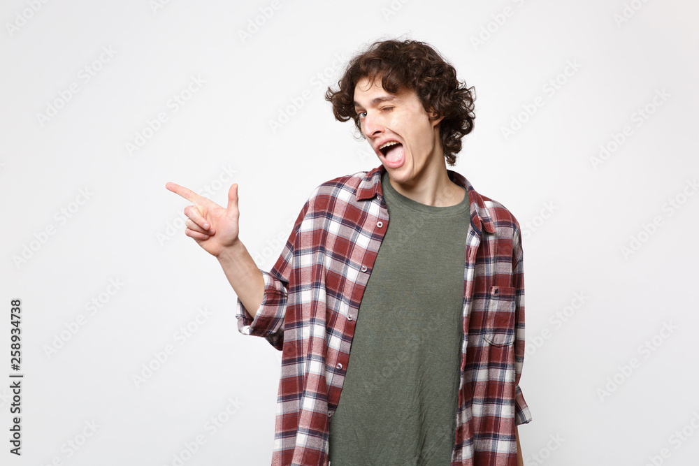 Portrait of funny blinking young man in casual clothes looking camera pointing index finger aside isolated on white background in studio. People sincere emotions lifestyle concept. Mock up copy space. Portrait of funny blinking young man in casual clothes looking camera pointing index finger aside isolated on white background in studio. People sincere emotions lifestyle concept. Mock up copy space.
