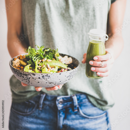 Healthy dinner or lunch. Woman in t-shirt and jeans standing and holding vegan superbowl or Buddha bowl with hummus, vegetable, salad, beans, couscous and avocado and smoothie in hands, square crop photo