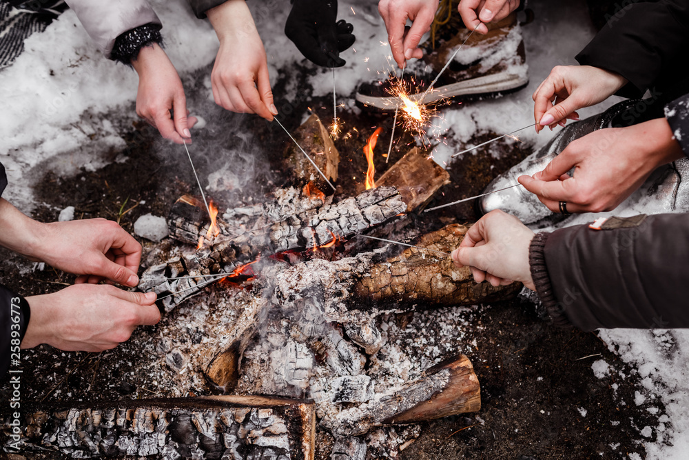 Obraz premium Young people sitting around campfire in the forest in winter and burning bengal lights. burn bonfire