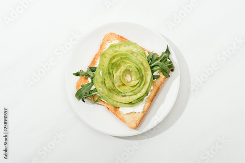Top view of toast with sliced avocado and arugula on plate on white surface
