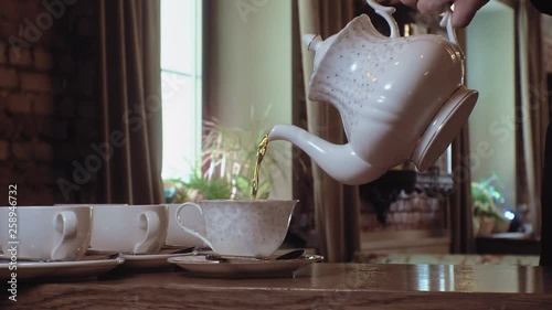A man pours tea from a kettle into a white cup on a wooden table.