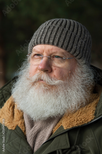 Stylish senior caucasian man in knitted cap and eyeglasses against dark natural winter background. Close up face of elderly hipster with splendid mustache and beard.