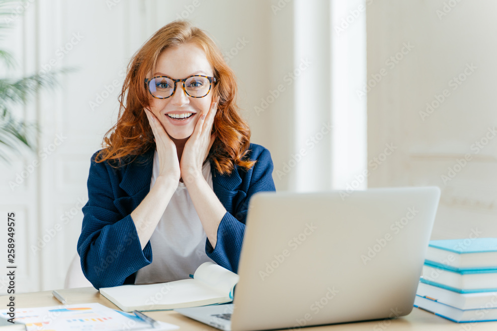 Positive redhead executive manager searches information on internet website, touches both cheeks, writes notes in notepad, uses modern laptop computer, dressed in formal clothes, checks organiser. Positive redhead executive manager searches information on internet website, touches both cheeks, writes notes in notepad, uses modern laptop computer, dressed in formal clothes, checks organiser.