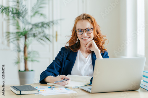 Photo of successful ginger female freelancer has remote work, watches webinar online on laptop computer, writes down information in notepad, smiles happily poses at workplace dressed in formal clothes photo