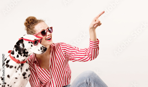 Happy girl and pet in red sunglasses. Young woman hugs dalmatian dog and points on right, isolated on white background. Check this out. Place for text