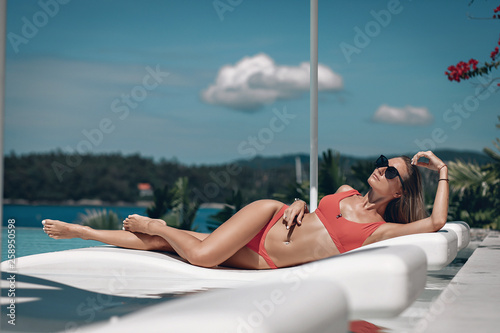 Side view of woman with perfect body, posing while sitting on open hotel terrace. Attractive smiling girl, wearing in red swimsuit, lying on white lounger near pool outside. Vacation. Phuket. Thailand © Semachkovsky 