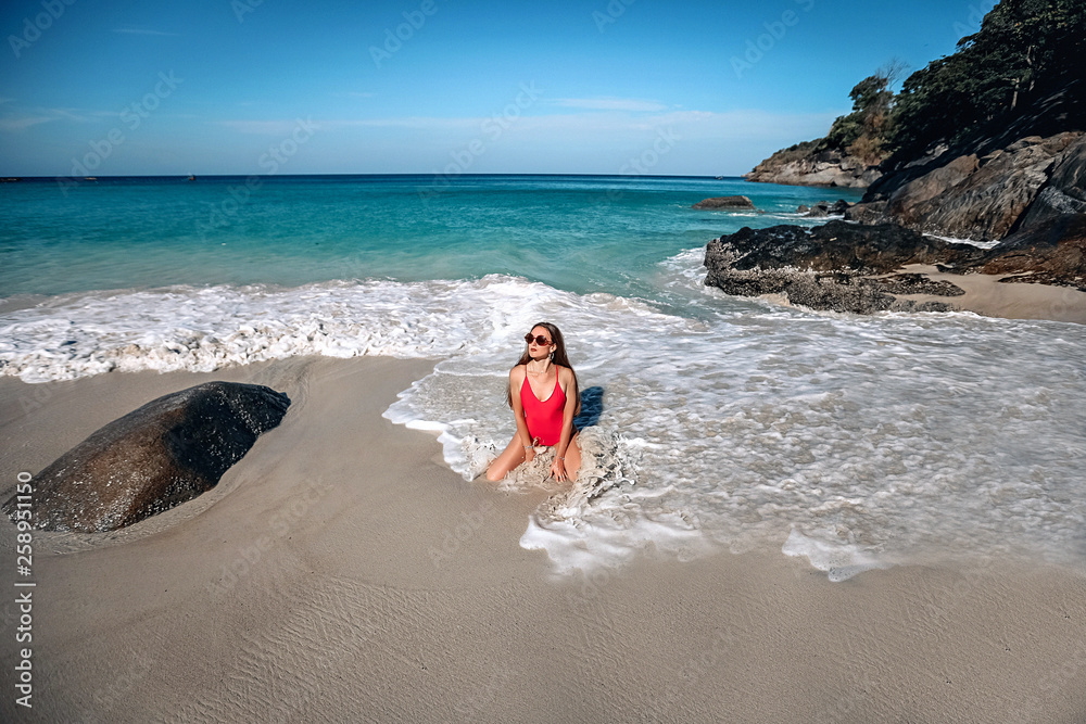 Young girl with a gorgeous body is resting on the beach with white sand near the ocean. Big sea waves. Beautiful sexy model in a red bathing suit and black sunglasses sunbathing. Phuket. Tailand Young girl with a gorgeous body is resting on the beach with white sand near the ocean. Big sea waves. Beautiful sexy model in a red bathing suit and black sunglasses sunbathing. Phuket. Tailand