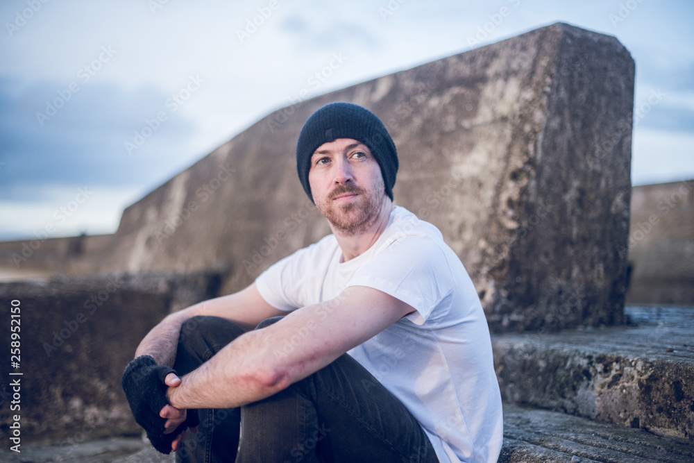 A young handsome man sitting alone and contemplating life by the quiet sea, surrounded by raw concrete sea defence wall. Mental health and depression road to recovery. A young handsome man sitting alone and contemplating life by the quiet sea, surrounded by raw concrete sea defence wall. Mental health and depression road to recovery.