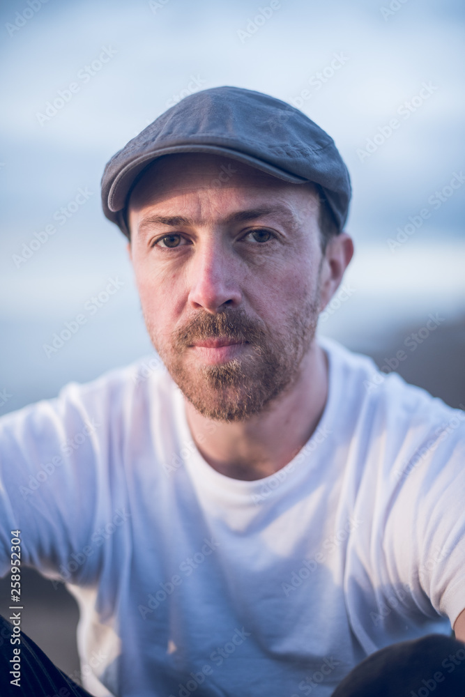 A portrait of young handsome man with stubble and a flat cap hat sitting alone and contemplating life by the quiet sea, surrounded by raw concrete sea defence wall. A portrait of young handsome man with stubble and a flat cap hat sitting alone and contemplating life by the quiet sea, surrounded by raw concrete sea defence wall.