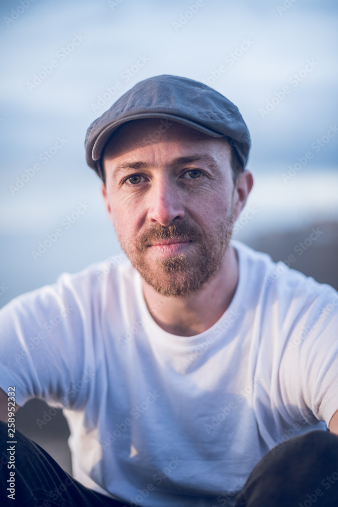 A portrait of young handsome man with stubble and a flat cap hat sitting alone and contemplating life by the quiet sea, surrounded by raw concrete sea defence wall. A portrait of young handsome man with stubble and a flat cap hat sitting alone and contemplating life by the quiet sea, surrounded by raw concrete sea defence wall.