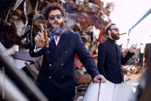 Charismatic bearded mens, wearings in stylish suit and sunglasses, smokes a cigar, on the old iron dump background