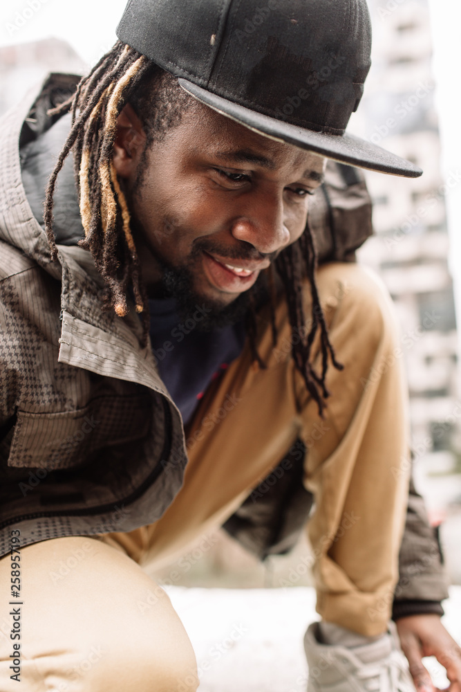Handsome African American with dreadlocks in brown trousers and a blue sweater in a black cap on his head against the background of houses, city Handsome African American with dreadlocks in brown trousers and a blue sweater in a black cap on his head against the background of houses, city