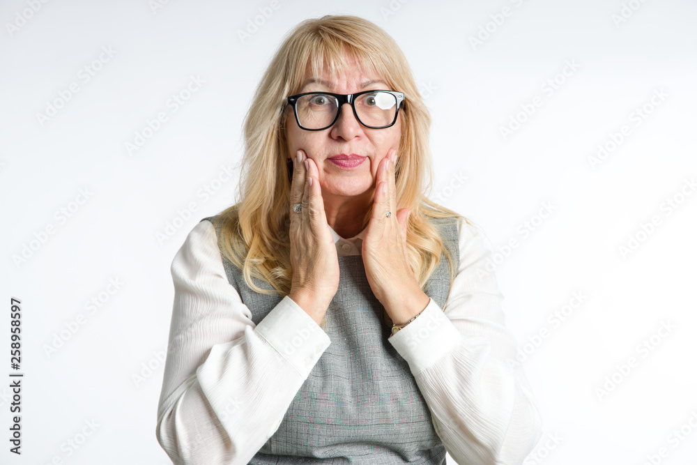Portrait of a mature woman with glasses. She examines her skin on her face while standing on a light background. Surprised woman. Portrait of a mature woman with glasses. She examines her skin on her face while standing on a light background. Surprised woman.