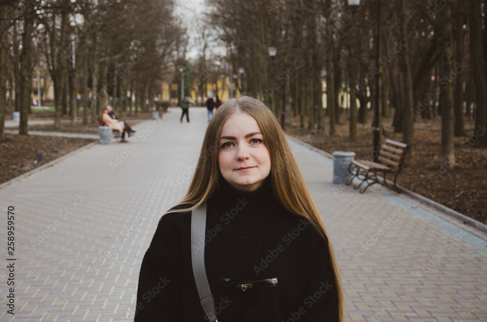 Portrait of a beautiful girl who walks along the alley in the park in a black jacket in the afternoon