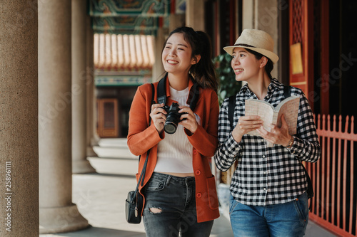two girls tourist visiting bangkok maining attractions in thailand. women friends travel walking in corridor hallway in chinese temple on sunny day in spring. backpackers smiling with camera and book photo