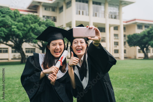 education graduation technology and people concept. group happy international asian students in mortar boards and bachelor gowns with diplomas having video phone call by smartphone outdoors thumb up