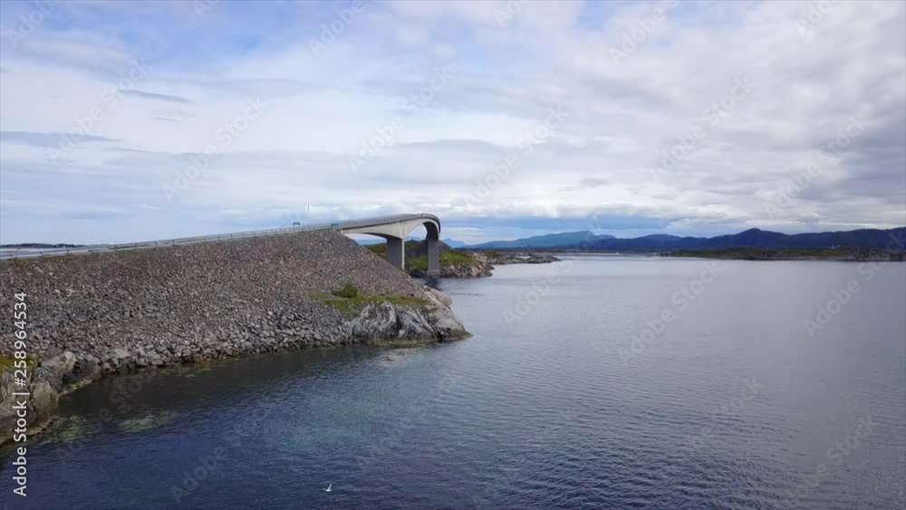 Aerial panning view of an arch bridge on the famous Norwegian Atlantic road, spanning a narrow strait in the Scandinavian fjords with scenic landscapes and dramatic clouds in the background Aerial panning view of an arch bridge on the famous Norwegian Atlantic road, spanning a narrow strait in the Scandinavian fjords with scenic landscapes and dramatic clouds in the background