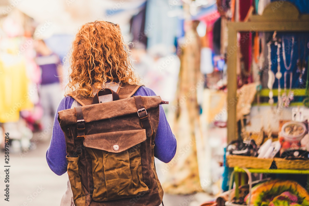 Obraz premium Blonde curly hair woman backpacker traveler viewed from rear at used market enjoying the shopping and alternative vacation - leather backpack view for travel concept