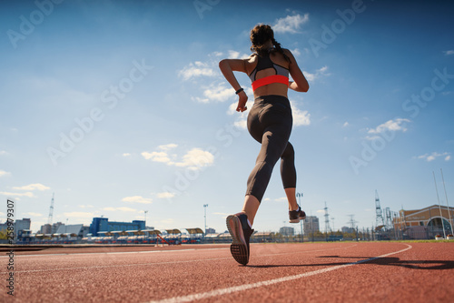 From below back view of athletic teen girl in sportswear with fitness tracker on her wrist running on track at stadium on summer day