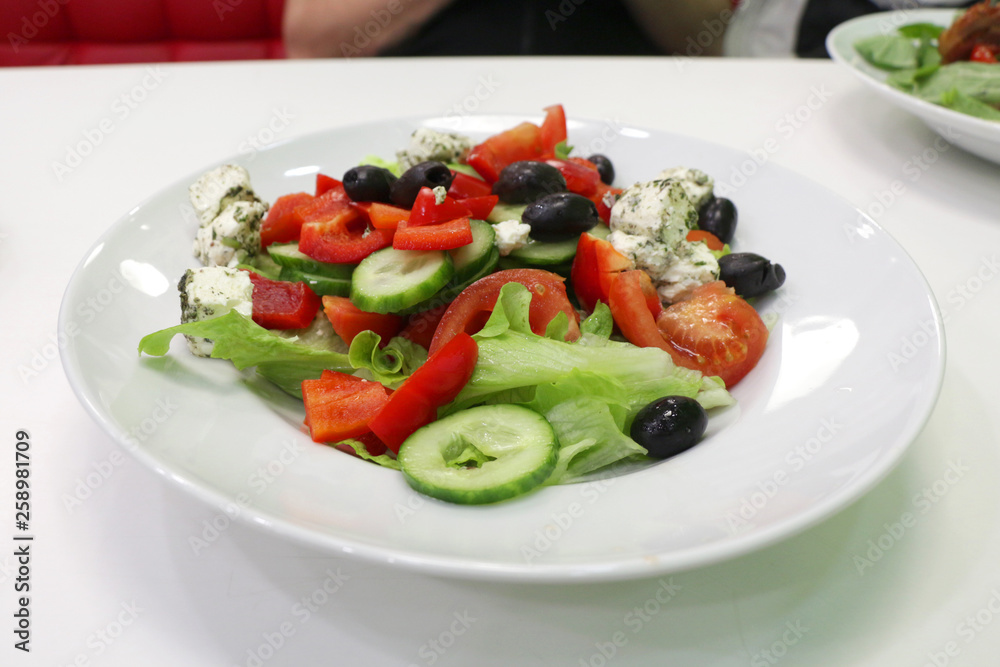 Greek salad of fresh cucumber, tomato, sweet pepper, lettuce, red onion, feta cheese and olives with olive oil in a white plate on a white table. View from above.