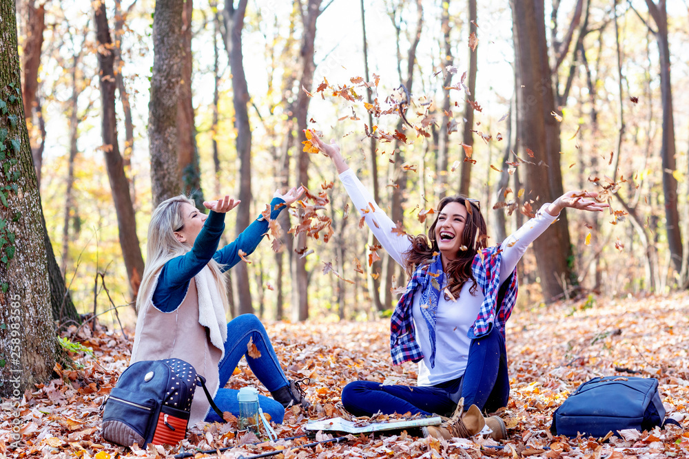Two girls relaxing in forest sitting on the ground and play witl leaves and smile Two girls relaxing in forest sitting on the ground and play witl leaves and smile