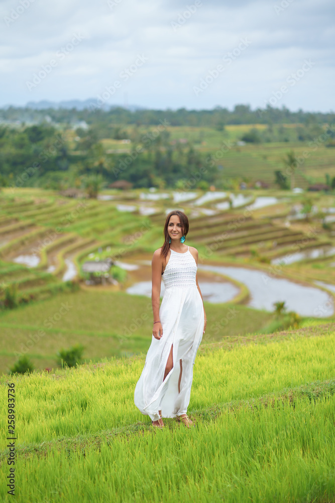 Tanned beautiful woman in white long dress smiling and posing against the background of rice fields Tanned beautiful woman in white long dress smiling and posing against the background of rice fields