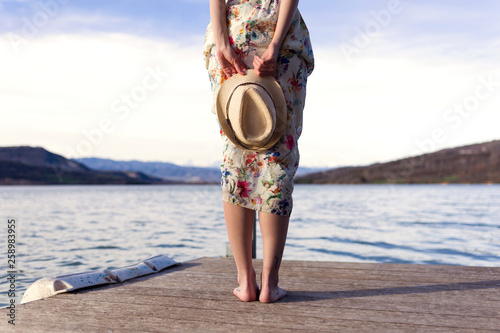 Back view of barefoot young woman standing on jetty holding summer hat, partial view photo