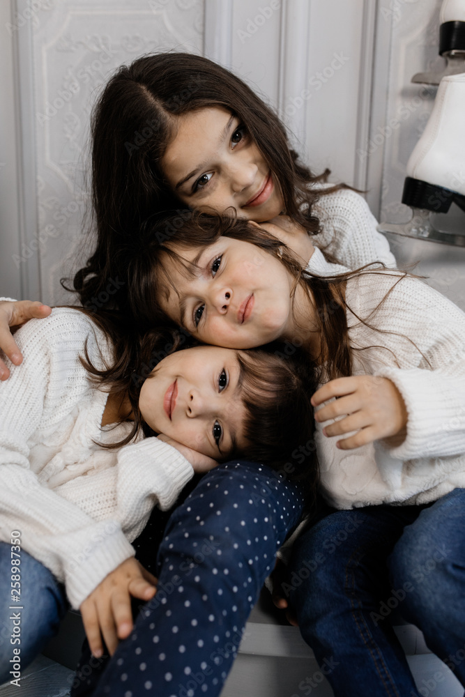 Three charming little sisters in white sweaters and blue jeans sit in a cozy room with Christmas decor Three charming little sisters in white sweaters and blue jeans sit in a cozy room with Christmas decor