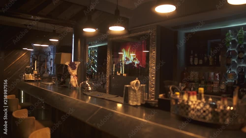 Beautiful and rich design of the bar in modern luxury restaurant. Comfortable chairs near bar counter, shelves with alcohol bottles and big electric fireplace on the wall in front of client sits