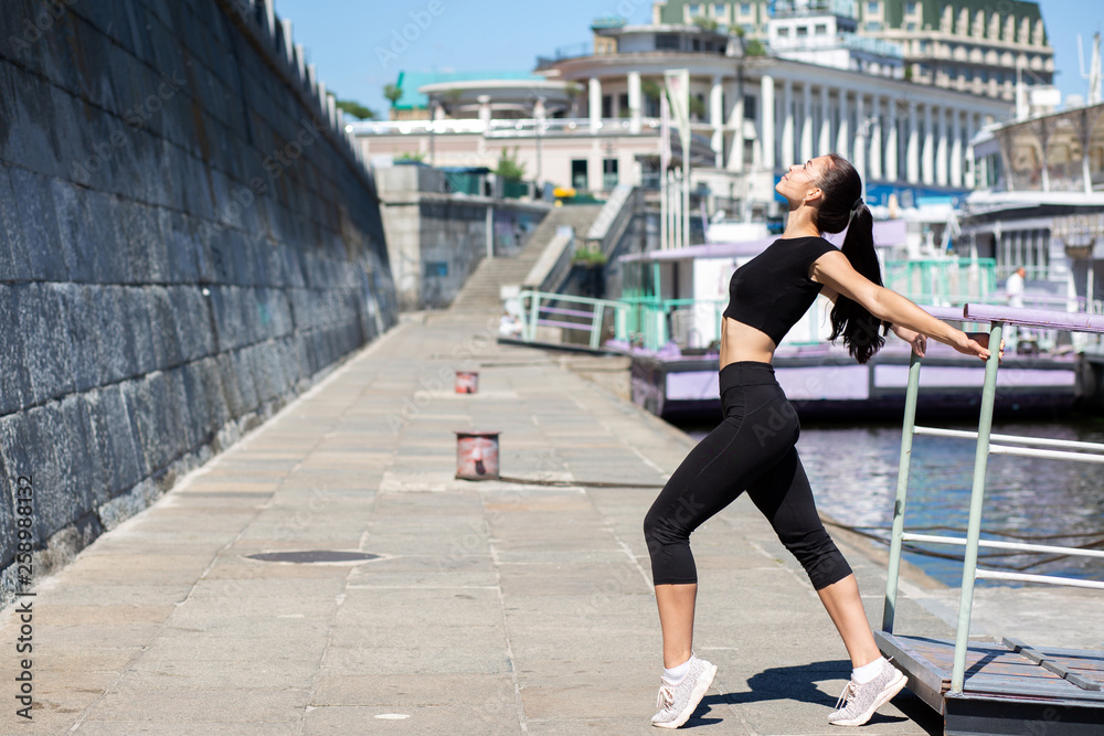 Slender brunette girl wearing sport apparel stretching at the city, near the river. Empty space