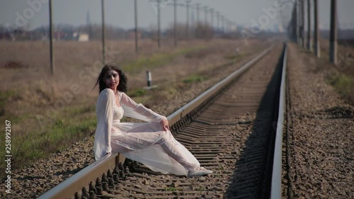 Amazing slim unusual beautiful girl in white translucent dress blown by wind and with wax makeup on face in form of bloody wound sits on railroad tracks in waiting