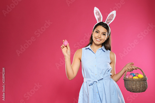 Beautiful woman in bunny ears headband holding basket with Easter eggs on color background, space for text