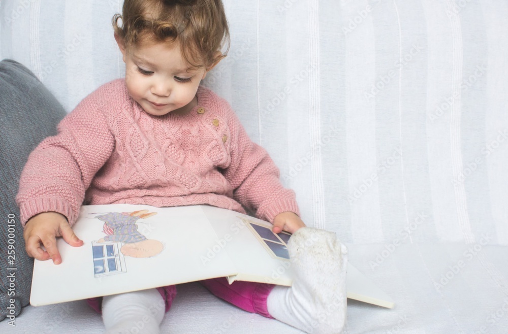 Little baby girl reading a picture book on a white sofa Little baby girl reading a picture book on a white sofa