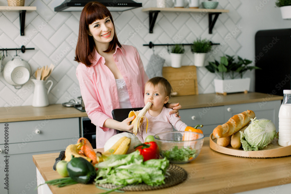 Cute young mother in pink shirt giving a banana to her baby girl sitting on the chair at the table with vegetables and fruits in the kitchen Cute young mother in pink shirt giving a banana to her baby girl sitting on the chair at the table with vegetables and fruits in the kitchen