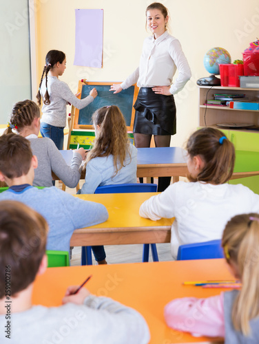 Positive female teacher standing at blackboard with schoolgirl during lesson in classroom
