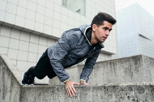 Portrait of sportsman use architecture for his calisthenic workout. He make push ups at concrete wall, in the background modern city buildings. Concept of focus on goals. © less.talk